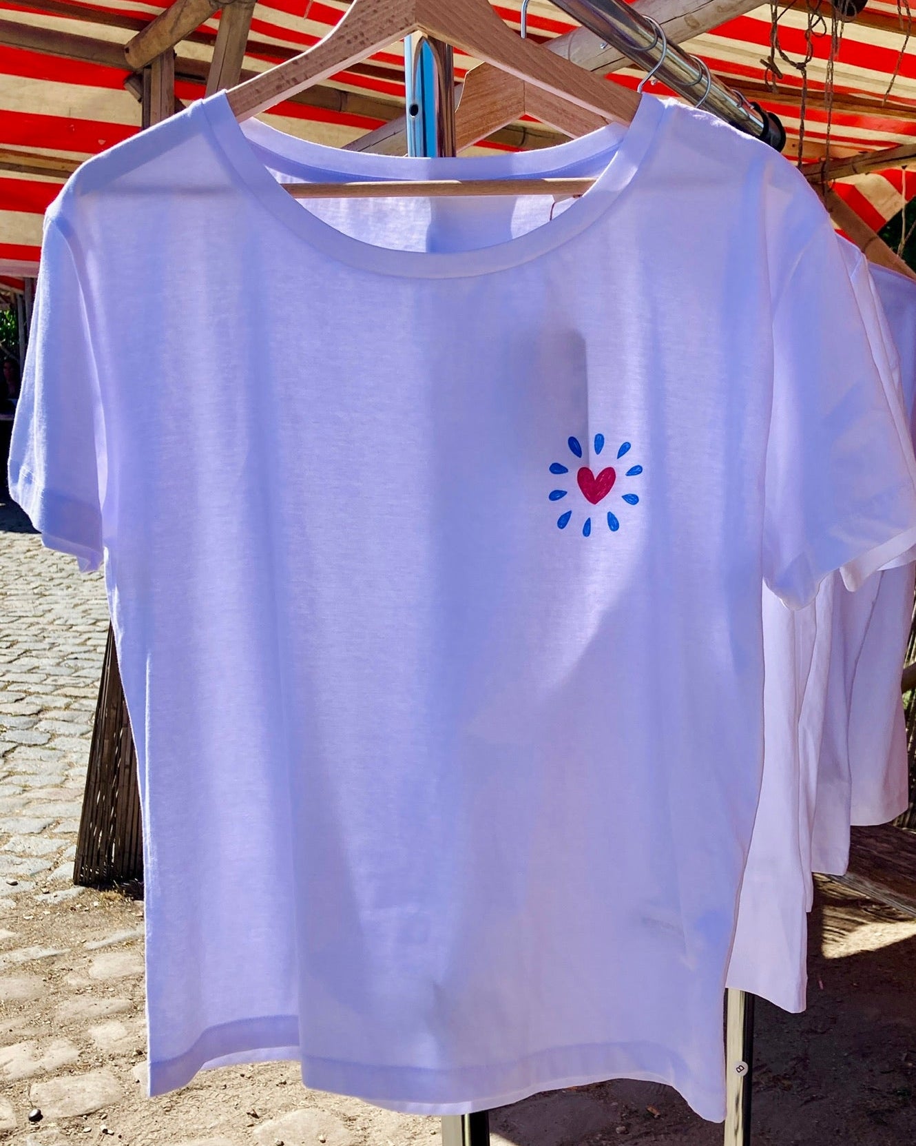 White t-shirt with a colorful heart design on a hanger against a beach background